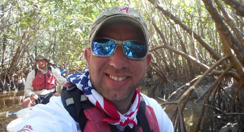 A person in a hat, sunglasses and an american flag bandana smiles from a canoe on an outward bound veterans expedition.